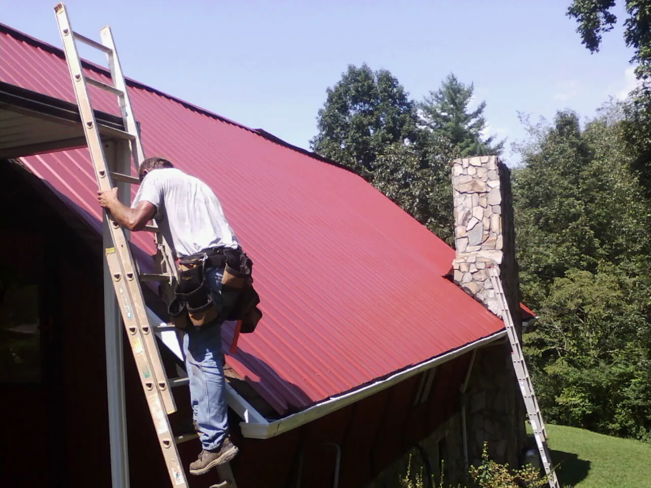 Roofer on a ladder beside a red metal roof and stone chimney — Soffit & Fascia Repair by Roofing Brothers in Eldorado Springs